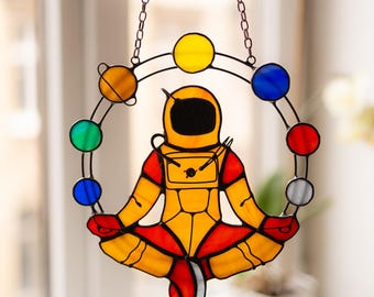 May include: A stained glass suncatcher featuring a yellow astronaut sitting in a lotus position, surrounded by a circular frame of small, colorful glass planets in shades of blue, green, orange, red, yellow, and white, suspended by a metal chain.