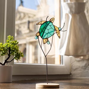 May include: A stained glass sea turtle sculpture with a green and blue shell and yellow accents. The turtle is perched on a wire branch with blue glass beads. The sculpture is on a wooden base.