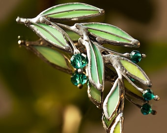 May include: A silver and green stained glass olive branch brooch with green crystal accents.