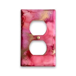 Könnte beinhalten: Eine rosafarbene und goldene Steckdosenabdeckung mit abstraktem Design. Die Abdeckung hat zwei Steckdosen und eine goldene Schraube in der Mitte.