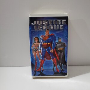 May include: A VHS tape featuring the title "JUSTICE LEAGUE" with illustrations of Superman, Wonder Woman, and Batman. The tape has a blue background with the Justice League logo at the top.
