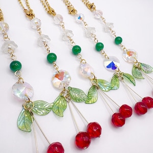 May include: Gold-toned chain necklaces with red cherry pendants. Each necklace features clear crystal beads, green beads, and green leaf accents. The cherries are a vibrant red color, and the overall design is delicate and feminine.
