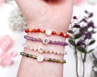 May include: Four beaded bracelets with gold initial charms. The bracelets are layered on a wrist and feature different colored beads, including red, purple, pink, and green.