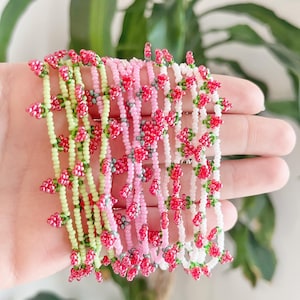 Peut inclure: Plusieurs bracelets de perles roses, blancs et vert clair. Chaque bracelet présente de petits motifs floraux perlés rouges et verts. Les bracelets sont présentés sur un fond doux.