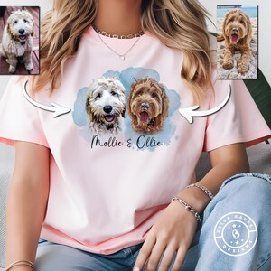 May include: A light pink short-sleeved t-shirt featuring a graphic print of two dogs with the names Mollie & Ollie written below them. The design includes a blue watercolor-style background and two smaller inset photos of the dogs above.