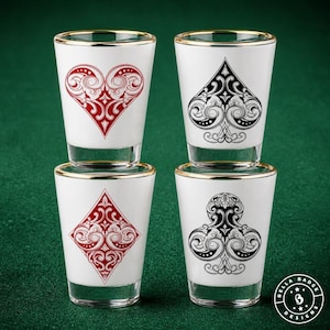 May include: A set of four white shot glasses with gold rims, each featuring a different playing card suit symbol in a decorative filigree style. The red heart and diamond symbols are on the left, while the black spade and club are on the right.