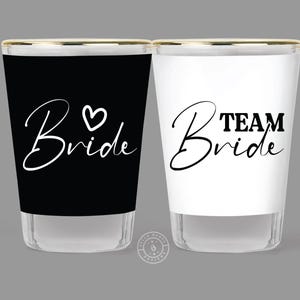 May include: Two shot glasses with gold rims. One is black with the text "Bride" and a heart. The other is white with the text "Team Bride".