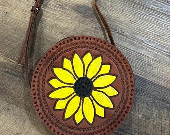 May include: Round brown leather crossbody bag with a hand-painted sunflower design. The sunflower has yellow petals and a black center. The bag has a brown leather strap and decorative stitching around the edge.