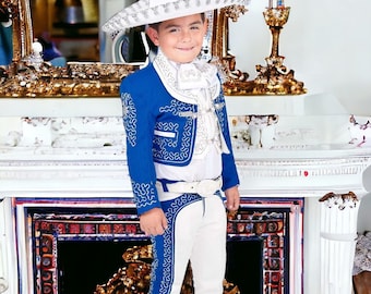 Puede incluir: Un niño pequeño con un traje de charro azul con bordados blancos, una camisa blanca y un sombrero de vaquero blanco. Está de pie frente a una chimenea blanca.