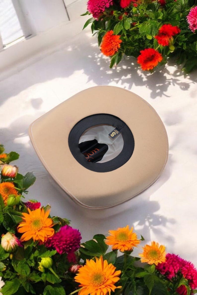 Peut inclure: Un chapeau de cowboy beige avec un bandeau noir et une petite &eacute;tiquette dor&eacute;e sur laquelle est inscrit "S". Le chapeau est entour&eacute; de fleurs orange et roses.