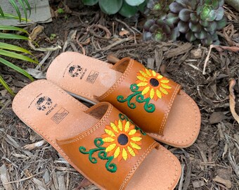May include: A pair of tan leather slide sandals featuring embroidered sunflower designs in yellow, orange, and green. The footbed displays a stamped logo with the text "MEXICAN ART" and a stylized face illustration, with visible stitching along the edges.