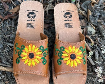May include: A pair of brown leather sandals with embroidered sunflowers. The sandals have a platform sole and are decorated with green vines and yellow and orange flowers. The sandals have the text "La Bella Mexicana" and "Mexican Art" printed on the back.