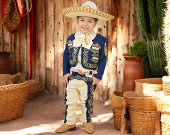 Puede incluir: Un niño pequeño vistiendo un traje de charro azul con bordados dorados y un sombrero de vaquero blanco. El traje incluye una chaqueta, pantalones y un chaleco. El niño sonríe y mira a la cámara.