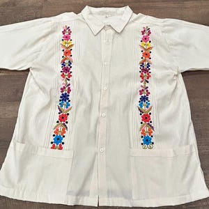 Peut inclure: Chemise en coton blanc à manches courtes et fermeture boutonnée. La chemise est ornée de motifs floraux brodés dans des tons de rose, bleu, vert, jaune et orange.