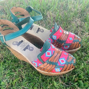 Peut inclure: Une paire de sandales en cuir vert et marron avec un motif tissé et un talon compensé. Les sandales ont un motif géométrique rose et bleu et le texte "MEXI LEATHER ART HECHO EN MEXICO" sur la semelle.