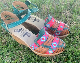 Puede incluir: Un par de sandalias de cuero verde y marrón con un diseño tejido y un tacón de cuña. Las sandalias tienen un patrón geométrico rosa y azul y el texto "MEXI LEATHER ART HECHO EN MEXICO" en la suela.