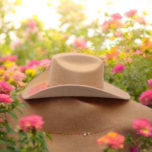 Peut inclure: Un chapeau de cowboy beige avec un large bord, reposant sur une surface brune entour&eacute;e de fleurs roses et jaunes.