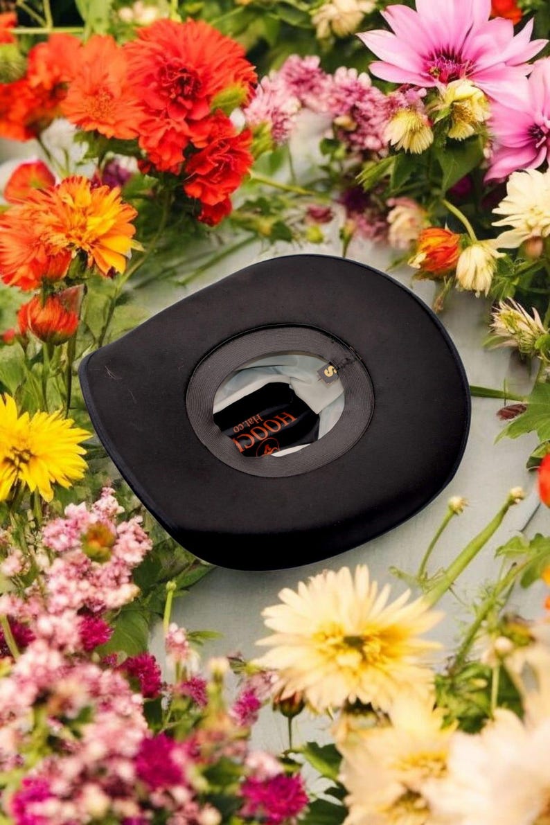 Puede incluir: Un sombrero vaquero de fieltro negro con una banda de cuero marr&oacute;n y las palabras "Hooey Hat Co." impresas en la banda. El sombrero est&aacute; rodeado de una variedad de flores coloridas.