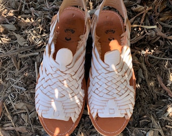 May include: A pair of white leather huarache sandals featuring a woven upper design, an open toe, and a heel strap. The tan leather insoles are stamped with the text Mi Bella Mexicana and the size 4, with white stitching along the outer edges.