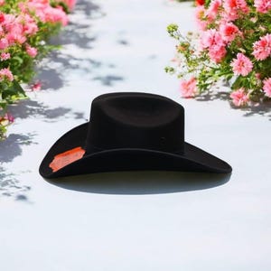 Puede incluir: Un sombrero vaquero de fieltro negro con un ala ancha. El sombrero est&aacute; sobre una superficie blanca.