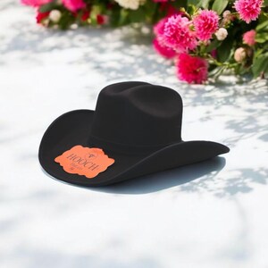 Puede incluir: Un sombrero vaquero de fieltro negro con un ala ancha y una peque&ntilde;a etiqueta naranja que dice "HOOCH" en blanco.