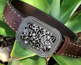 May include: A brown leather belt with a decorative silver buckle. The buckle features an ornate floral design with clear crystal accents. The belt has small silver studs along the edges.