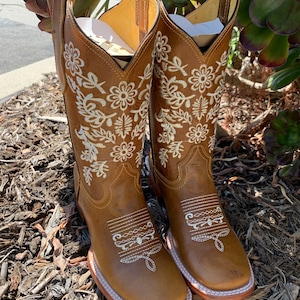 May include: A pair of brown leather cowboy boots with white floral embroidery. The boots have a pointed toe and a low heel.