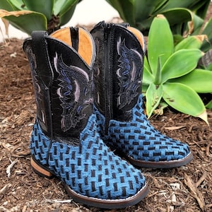 May include: A pair of children's cowboy boots. The boots have black leather uppers with embroidered designs and blue woven fabric on the foot. The boots are set on a bed of mulch with green plants in the background.