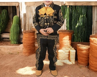 Puede incluir: Un niño pequeño vistiendo un traje de charro negro y dorado con detalles bordados. El traje incluye una chaqueta, pantalones y una corbata de moño. El niño está de pie frente a un fondo rústico con cactus.
