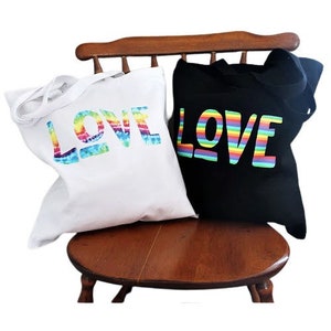 May include: Two tote bags with the word "LOVE" printed on them. The bag on the left has a rainbow tie-dye design and the bag on the right has a rainbow stripe design.