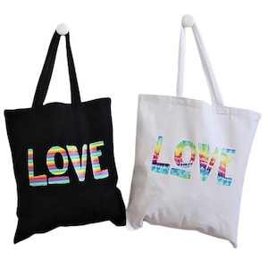 May include: Two tote bags with the word "LOVE" printed on them. One bag is black with rainbow stripes in the letters. The other bag is white with rainbow tie-dye in the letters.