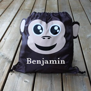 May include: A brown drawstring bag with a monkey face on the front. The monkey has big eyes, a smile, and a brown nose. The bag has the name "Benjamin" printed in white letters on the front.