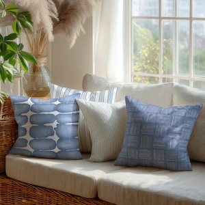 May include: A collection of decorative throw pillows in shades of blue and white arranged on a beige sofa. The pillows feature various patterns, including watercolor-style shapes, stripes, and a woven texture. The scene is set near a window with natural light.