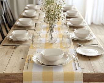 May include: A yellow and white checkered table runner with a wooden table and white plates, silverware, and glasses set for a dinner party. A vase with white flowers is in the center of the table.