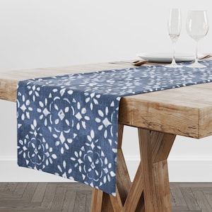 May include: A blue table runner with a white floral pattern is displayed on a wooden table. The runner is made of fabric and has a repeating design of white shapes against a blue background. The table is set with plates, silverware, and wine glasses.
