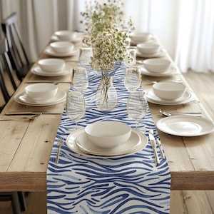 May include: A blue and white striped table runner with a watercolor-like design. The runner is laid out on a wooden table with white plates and glasses.