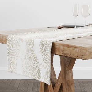 May include: A white table runner with a beige paisley pattern. The runner is laid on a wooden table with a white plate and silverware.
