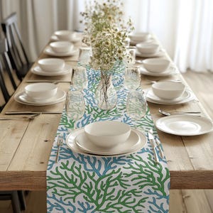 Könnte beinhalten: Ein weißer Tischläufer mit einem grünen und blauen Korallenriff-Design. Der Läufer liegt auf einem Holztisch mit weißen Tellern, Schalen und Gläsern.