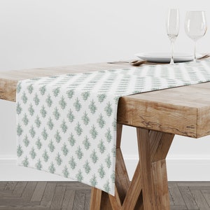 Op de afbeelding: Een witte tafelrunner met een groen bladmotief. De runner ligt op een houten tafel met twee wijnglazen en een bord.