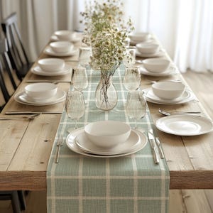 Op de afbeelding: Een lichtgroene en witte geruite tafelrunner met een vaas met witte bloemen en een set witte borden en kommen op een houten tafel.