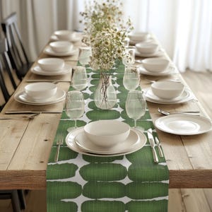 Peut inclure: Un chemin de table vert et blanc avec un motif abstrait d'ovales verts. Le chemin de table est posé sur une table en bois avec des assiettes blanches, des verres et des couverts disposés pour un dîner.
