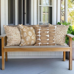 Könnte beinhalten: Vier dekorative Zierkissen mit verschiedenen Mustern liegen auf einer Holzbank. Die Kissen zeigen Designs in Weiß und Beige, darunter geometrische Formen, Blättermotive und abstrakte Formen. Die Bank steht auf einer Veranda mit weißer Tür und Fenster.