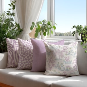 Op de afbeelding: Een verzameling decoratieve sierkussens in tinten lavendel en wit, gerangschikt op een witte bank. De kussens hebben verschillende patronen, waaronder een zebraprint, stippen en een landschapsontwerp. De scène speelt zich af bij een raam met planten.