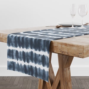 May include: A blue and white tie-dye patterned table runner. The runner is draped over a wooden table, with the design featuring horizontal stripes of blue and white. The table is set with plates, silverware, and wine glasses.