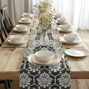 Peut inclure: Un chemin de table noir et blanc à motif damassé avec un motif floral blanc. Le chemin de table est posé sur une table en bois avec des assiettes et des bols blancs, des couverts et un vase de fleurs blanches.