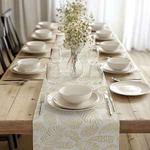 Könnte beinhalten: Ein weißer Tischläufer mit einem goldenen Blattmotiv. Der Läufer liegt auf einem Holztisch mit weißen Tellern, Schalen und Gläsern, die für ein Abendessen gedeckt sind. Ein Strauß weißer Blumen steht in einer durchsichtigen Glasvase in der Mitte des Tisches.
