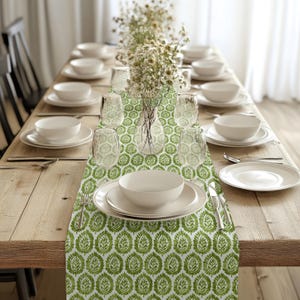 May include: A green and white table runner with a leaf pattern. The runner is laid out on a wooden table with white plates, bowls, and glasses. A vase with white flowers is in the centre of the table.