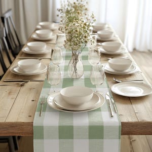 May include: A green and white checkered table runner with a vase of white flowers and a place setting with a white bowl and plate.