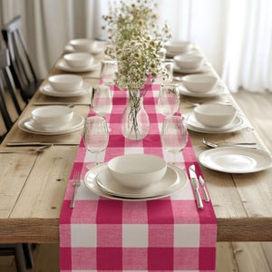 Puede incluir: Un camino de mesa a cuadros rosa y blanco con fondo blanco y cuadrados rosas. El camino de mesa está colocado sobre una mesa de madera con platos blancos, vasos y cubiertos dispuestos para una cena.