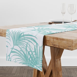May include: A teal and white table runner with a tropical leaf design. The runner is laid on a wooden table with two wine glasses and a plate.